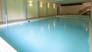 Ein Hallenbad mit glasklarer Wasseroberfläche, einer Treppe zu einer erhöhten Ebene und einigen Liegestühlen in der Nähe des Beckens.