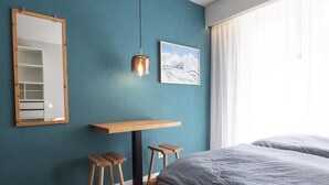 Ein Schlafzimmer mit einem Bett, einem Schreibtisch, zwei Hocker, einem Spiegel, einer Pendelleuchte und einem gerahmten Bild an der Wand.