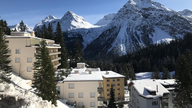 Ein Bergurlaubsort mit schneebedeckten Gebäuden, darunter ein mehrstöckiges Hotel und ein kleineres Bauwerk, vor einer Kulisse schneebedeckter Berggipfel.