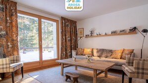 Ein gemütliches Wohnzimmer mit einer Couch, Sesseln, einem Couchtisch und einem großen Fenster mit Vorhängen.