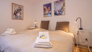 Ein Hotelzimmer mit einem Bett, zwei Kissen, einem Nachttisch mit Lampe, einer Wand mit zwei Postern und gefalteten Handtüchern auf dem Bett.