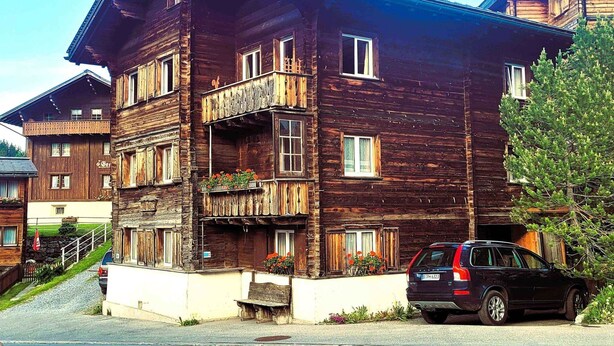Ein mehrstöckiges Holzgebäude mit Balkonen, ein Auto vor der Fassade und ein Baum zur Rechten.
