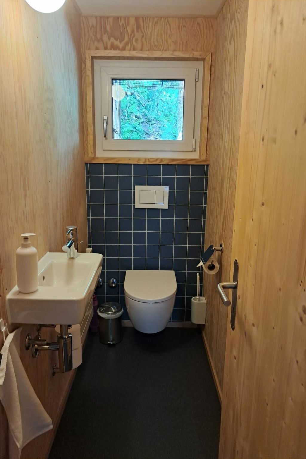 A compact bathroom with a wooden wall, a window above the toilet, a sink with a soap dispenser, and a toilet paper holder.