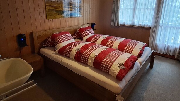 A double bed with checkered bedding, a wooden headboard, a mounted painting, a bedside lamp, a window with sheer curtains, and a wooden floor. - Chalet Suveran 1 Appartement/Fewo, Bad, WC, 6-Bettwohnung Suveran 1 by Interhome (Arosa)