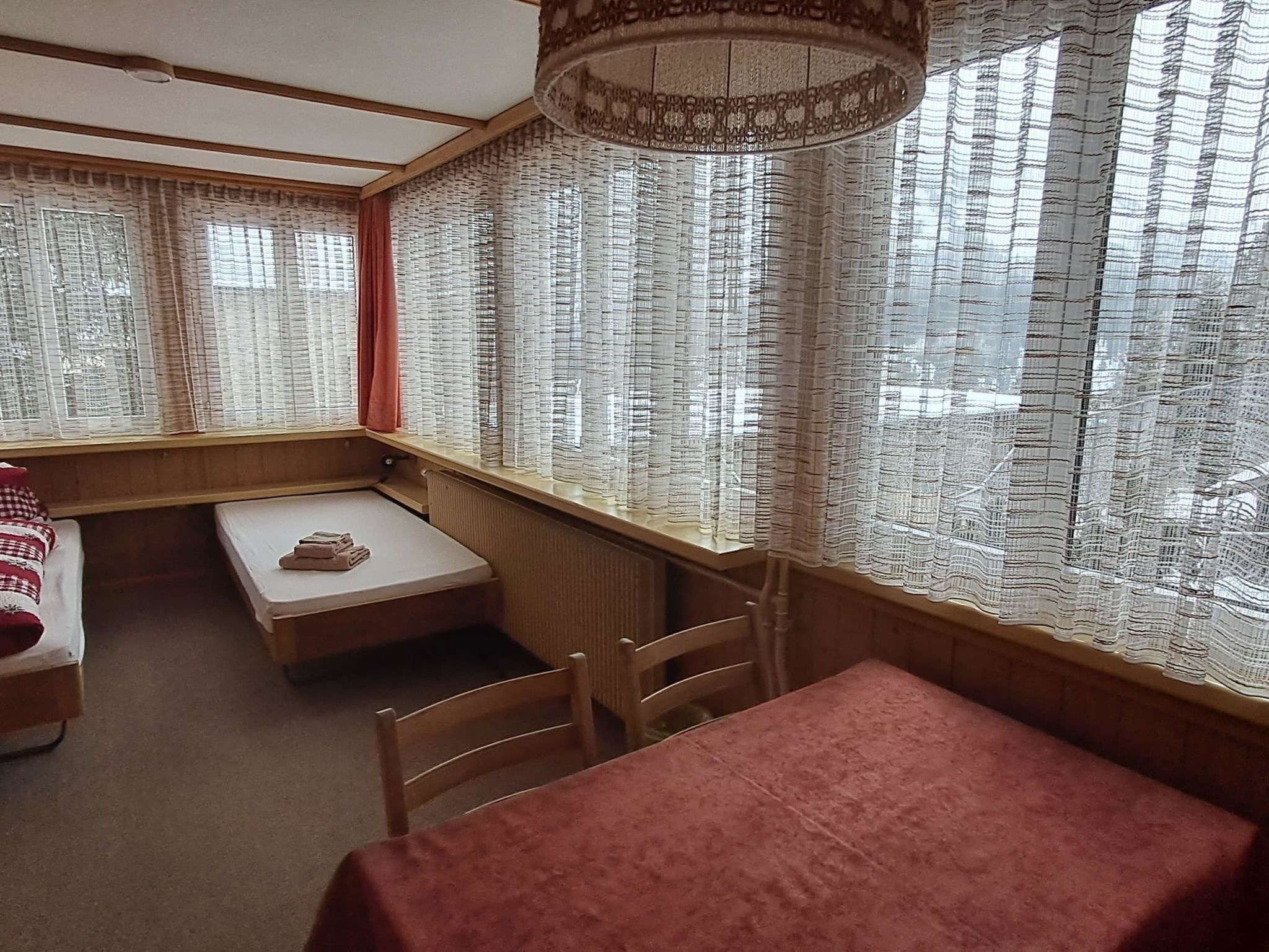 Ein Zimmer mit einem Bett, einem kleinen Tisch, einer Bank und einem Fenster mit gemusterten Vorhängen.