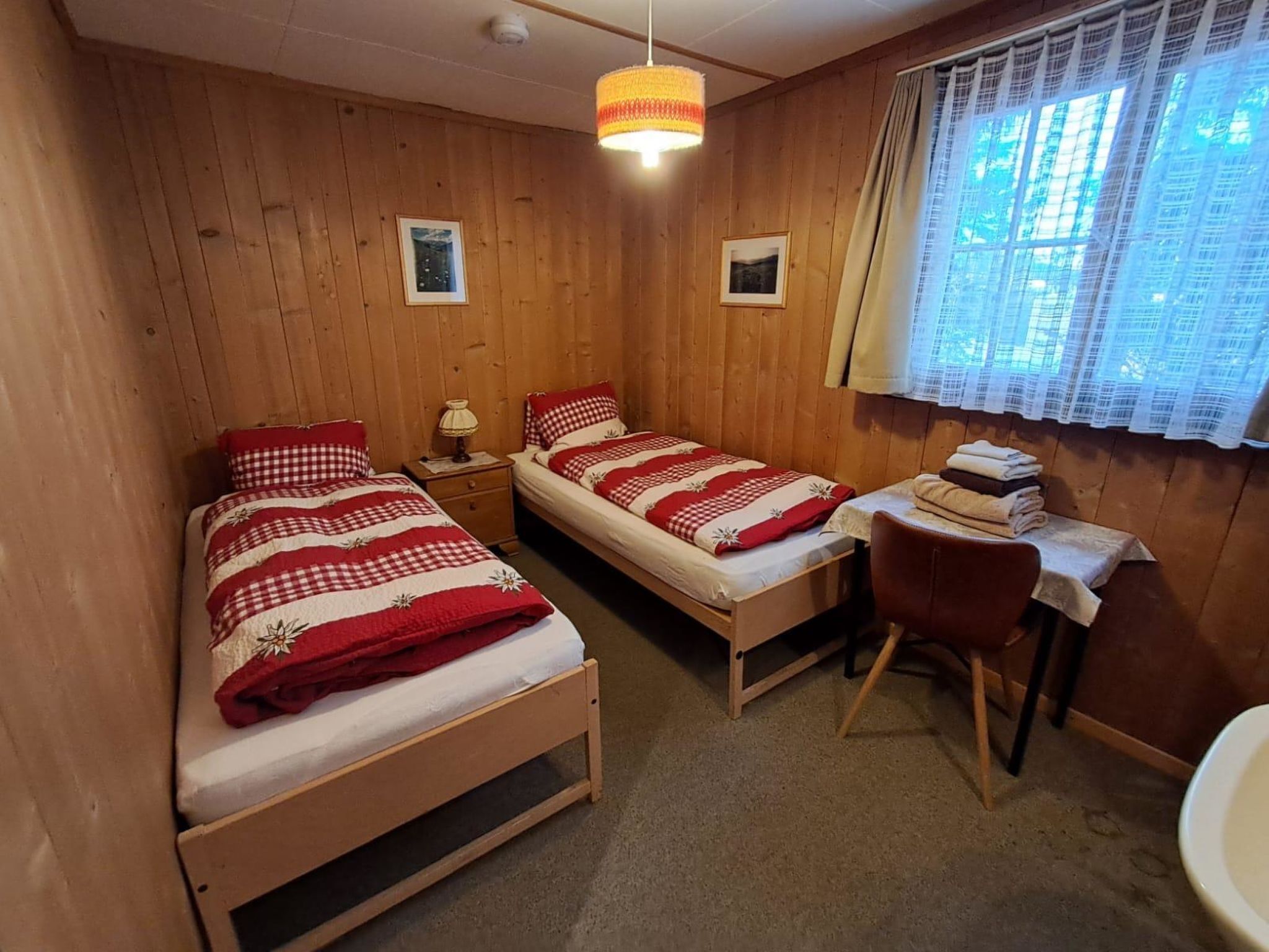 A room with two wooden beds, a desk with a chair, a hanging light fixture, and a window with curtains.