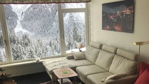 A cozy living room with a sofa, a small table, a lamp, and a view of snowy mountains through large windows. - Ritschis Home Appartement/Fewo, Dusche, WC, 4-Bettwohnung Ritschis Home by Interhome (Arosa)
