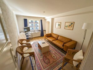Ein Wohnzimmer mit braunem Sofa, Korbsesseln, Couchtisch, Fernseher und Heizkörper.