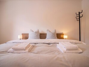 Ein ordentlich bezogenes Bett mit Kissen und gefalteten Handtüchern, eine Nachttischlampe und ein Kleiderständer.