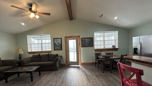 A spacious living room with a ceiling fan, a dining area with wooden chairs, a kitchen with a refrigerator, and large windows allowing natural light.