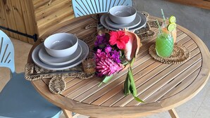 Een ronde houten tafel met een geweven placemat, twee witte kommen, een glas met een groen drankje en een bloemenboeket.