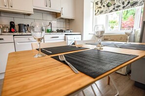 A dining table with two wine glasses and cutlery, a kitchen in the background, and a window with a view of greenery. - Howard Road Sleeps 7 -5 Bedrooms | Parking |Garden (Bristol)