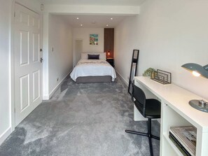 A narrow hallway leading to a bed with a white headboard and a single pillow, a desk with a chair, a mirror, and a small plant on the desk. - Howard Road Sleeps 7 -5 Bedrooms | Parking |Garden (Bristol)