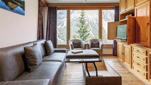 Un soggiorno moderno con divano, poltrone, tavolino, mobili in legno, televisione e un'ampia finestra vista montagna.