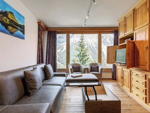 Ein modernes Wohnzimmer mit einer Couch, Sesseln, einem Couchtisch, Holzschränken, einem Fernseher und einem großen Fenster mit Blick auf die Berge.