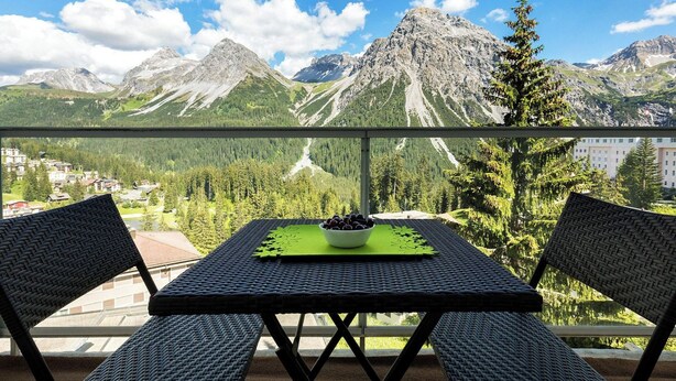Un balcone con sedie in vimini e un tavolo, con vista su un paesaggio montuoso e un villaggio sottostante.