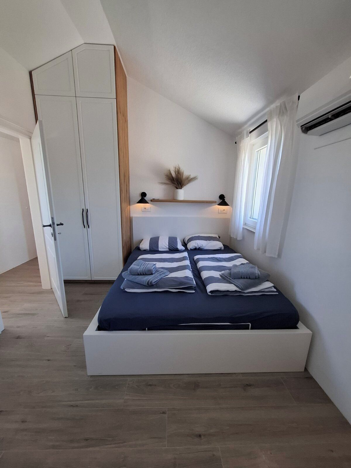 Een slaapkamer met een bed, een garderobe, een plankje met een plant en een raam met gordijnen.