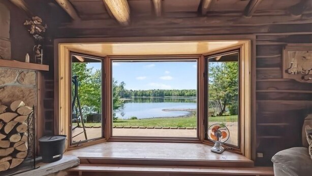 Una cabaña con un amplio ventanal con vista a un lago, una chimenea y un ventilador en el alféizar de la ventana.