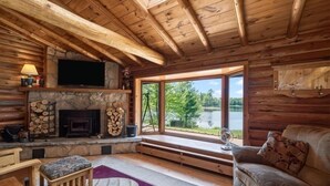 Interior de una cabaña de madera con chimenea de piedra, un televisor montado encima, un sofá y un ventanal con vistas a un lago.