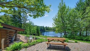 Una cabaña de madera con una mesa y sillas de picnic, un patio empedrado y vistas a un lago.