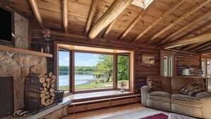 Interior de una cabaña de troncos con chimenea de piedra, un sofá, un ventanal grande con vista a un lago y techo de madera.