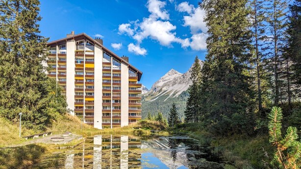 A multi-story hotel with balconies, a lake in front, and mountains in the background. - Rothornblick 11 by Arosa Holiday Appartement/Fewo, Bad, WC Rothornblick 11 by Interhome (Arosa)