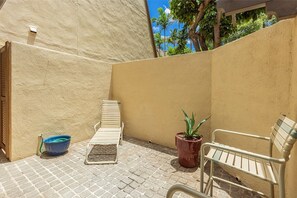 A patio area with a brick floor, two metal benches, a potted plant, and a blue bucket. - Maui Kamaole B106 1 Bedroom Apts (Kihei)