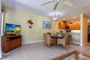 A living room with a TV, dining table, chairs, a ceiling fan, and a kitchen area. - Maui Kamaole B106 1 Bedroom Apts (Kihei)