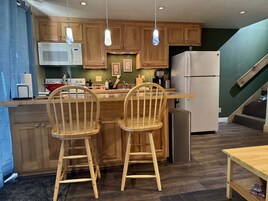 Una cocina con armarios de madera, un microondas, una estufa, un refrigerador y dos sillas de madera.