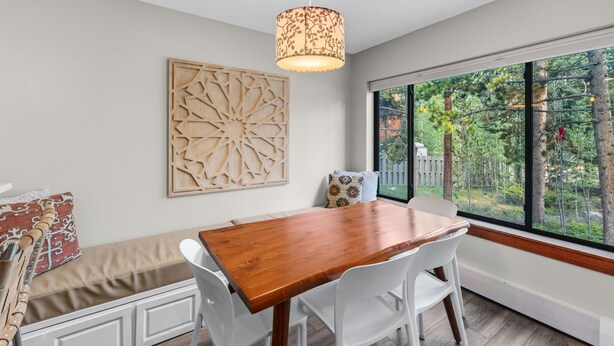 A dining area with a wooden table and white chairs, a decorative wall piece, and a view of trees through large windows. - Quiet Keystone Condo Near Slopes, Lake Dillon, Pool and Hot Tub (Keystone)