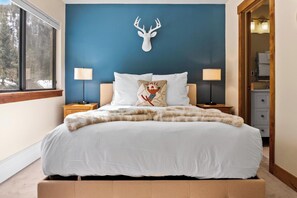 A bedroom with a large bed, two bedside tables with lamps, a deer head wall decoration, and a window with a view of trees. - Quiet Keystone Condo Near Slopes, Lake Dillon, Pool and Hot Tub (Keystone)