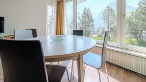 A dining area with a round table, chairs, a glass bowl, a hanging light, and a large window with a mountain view. - Viktoria A12 by Arosa Vacations Appartement/Fewo, Dusche, WC, 1 Schlafraum by Interhome (Arosa)