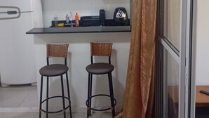 Une cuisine équipée d’un réfrigérateur, d’un four à micro-ondes et d’une machine à café sur le plan de travail. On y trouve également deux chaises et un rideau près de la fenêtre.