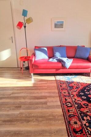Ein rotes Sofa mit blauen Kissen, ein gemustertes Teppich, ein kleiner roter Tisch und eine Stehlampe mit farbigen Lampenschirmen.