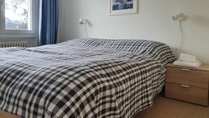 Et soveværelse med seng, et natbord, en lampe, et vindue med gardiner og et billede i ramme på væggen.