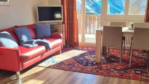 Et værelse med en rød sofa, et fjernsyn, et spisebord med stole og udsigt til sneklædte bjerge gennem vinduet.