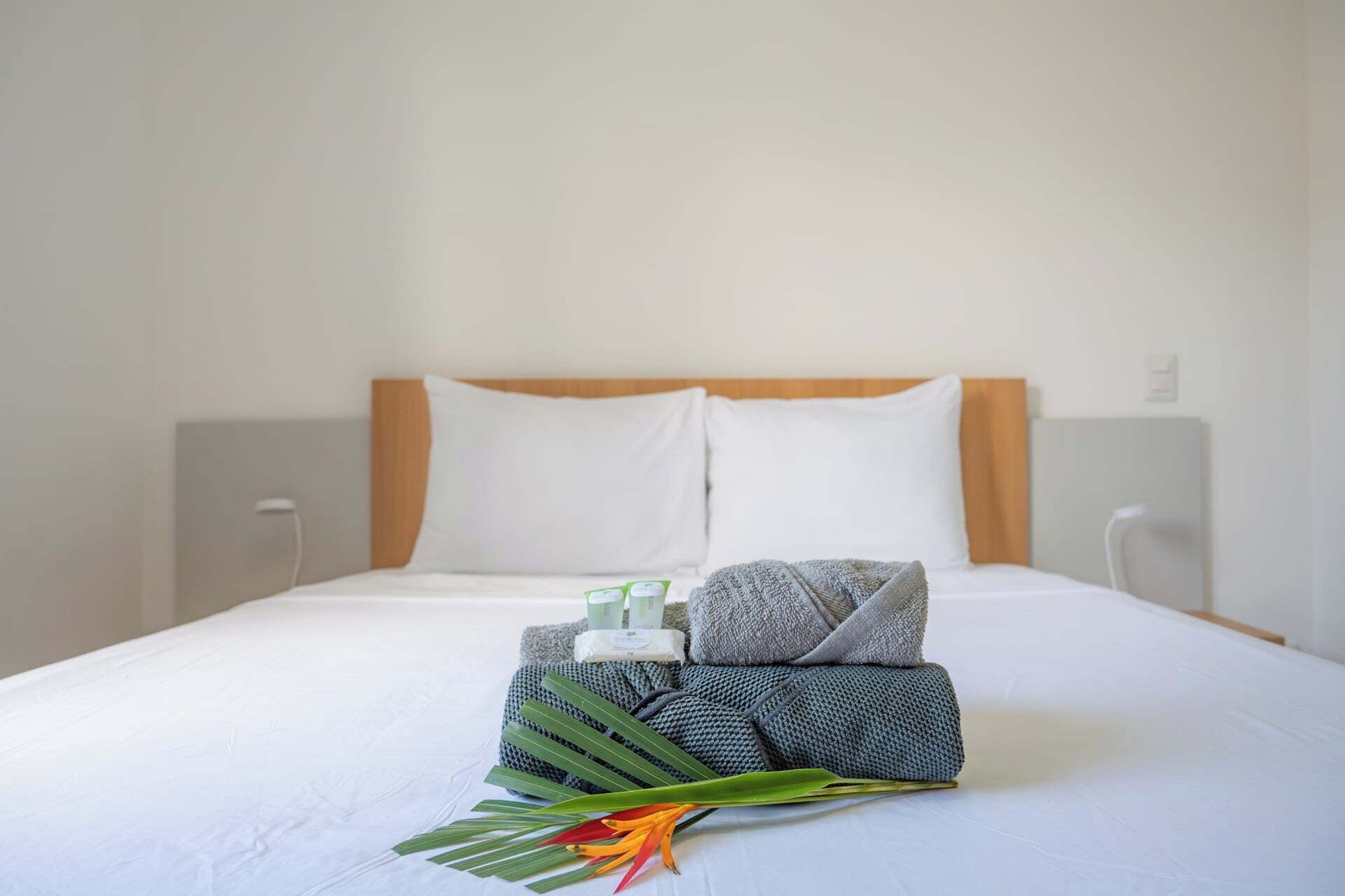 A neatly made bed with white bedding, a grey and black patterned towel, a green plant, and a bottle of lotion.