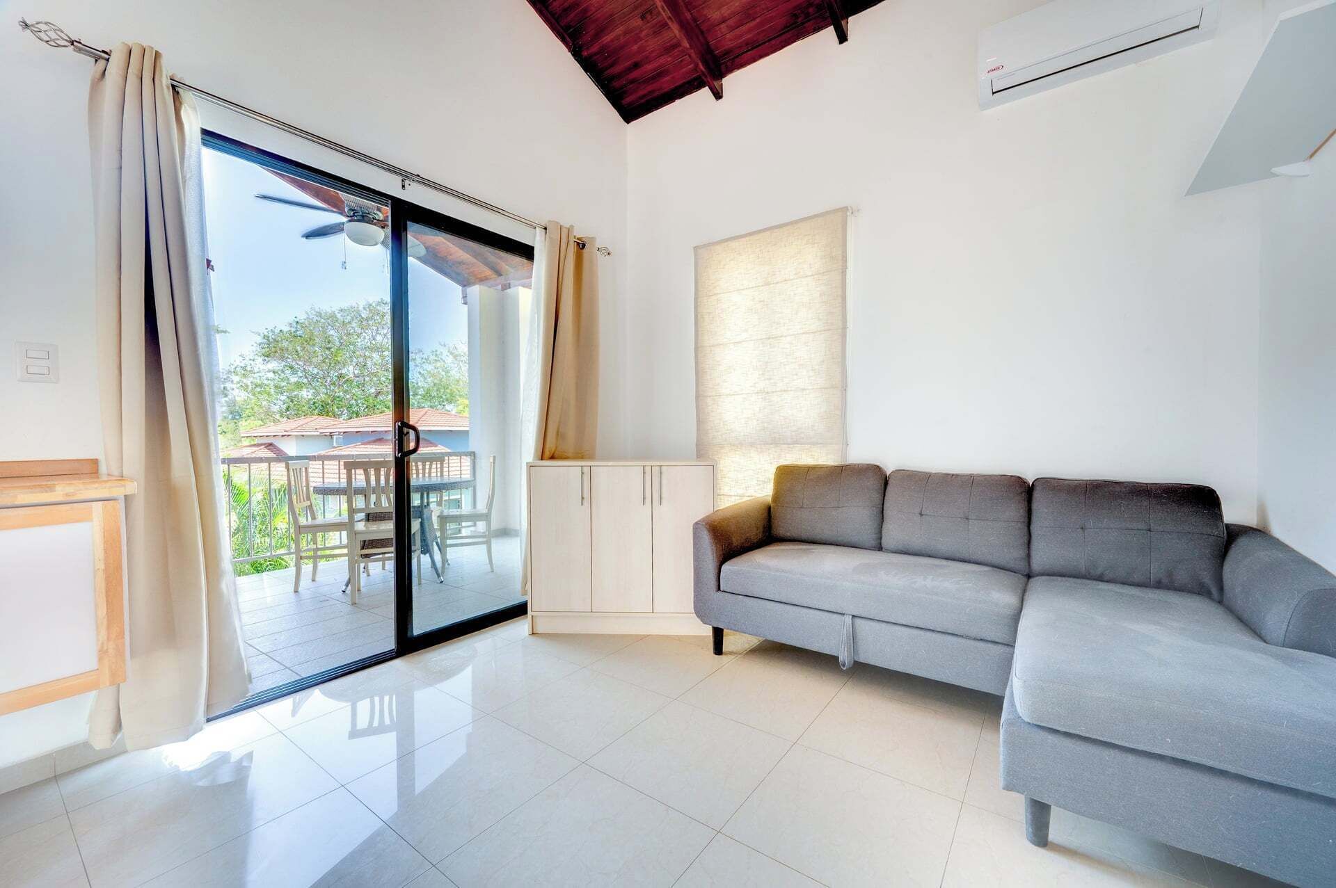 A living room with a grey sofa, a sliding glass door leading to an outdoor area, a ceiling fan, and an air conditioning unit.