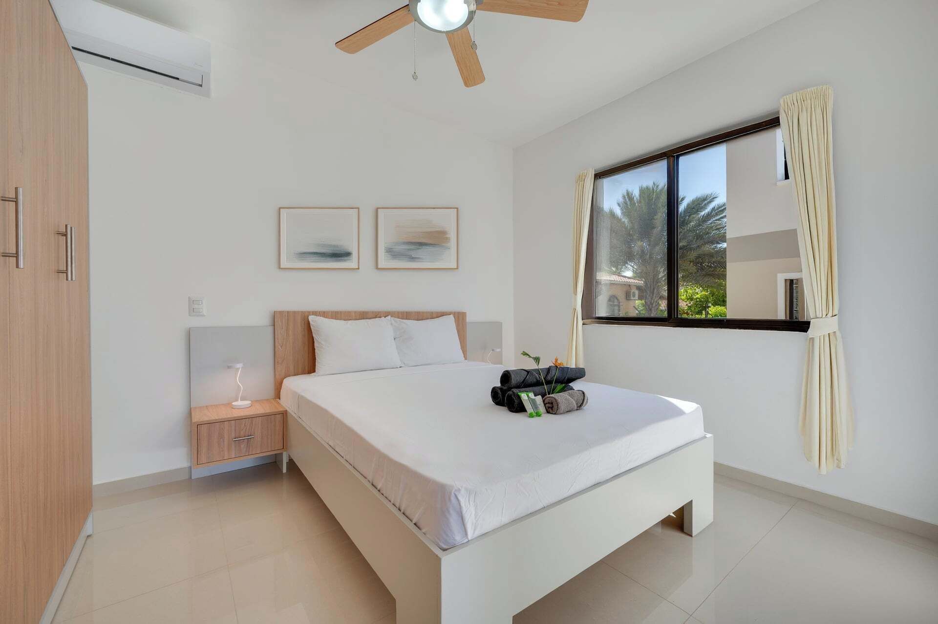 A bedroom with a bed, a nightstand, a ceiling fan, a window with curtains, and two framed pictures on the wall.