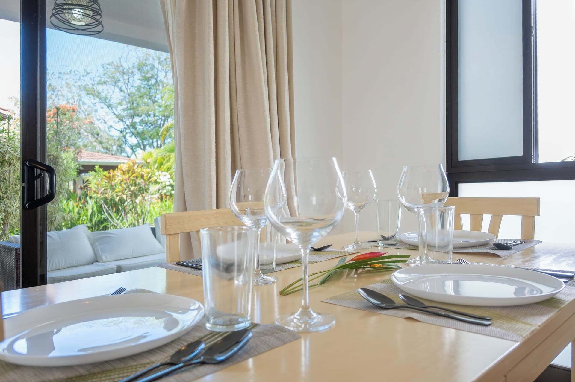 Mesa de comedor servida con platos, cubiertos y copas de vino, con vista a follaje verde al exterior.