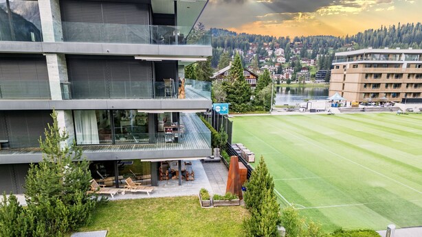 A modern building with a glass facade, outdoor seating, and a view of a green field and distant buildings. - Eden 3 by Arosa Holiday Appartement/Fewo, Bad, WC, 6-Bettwohnung by Interhome (Arosa)