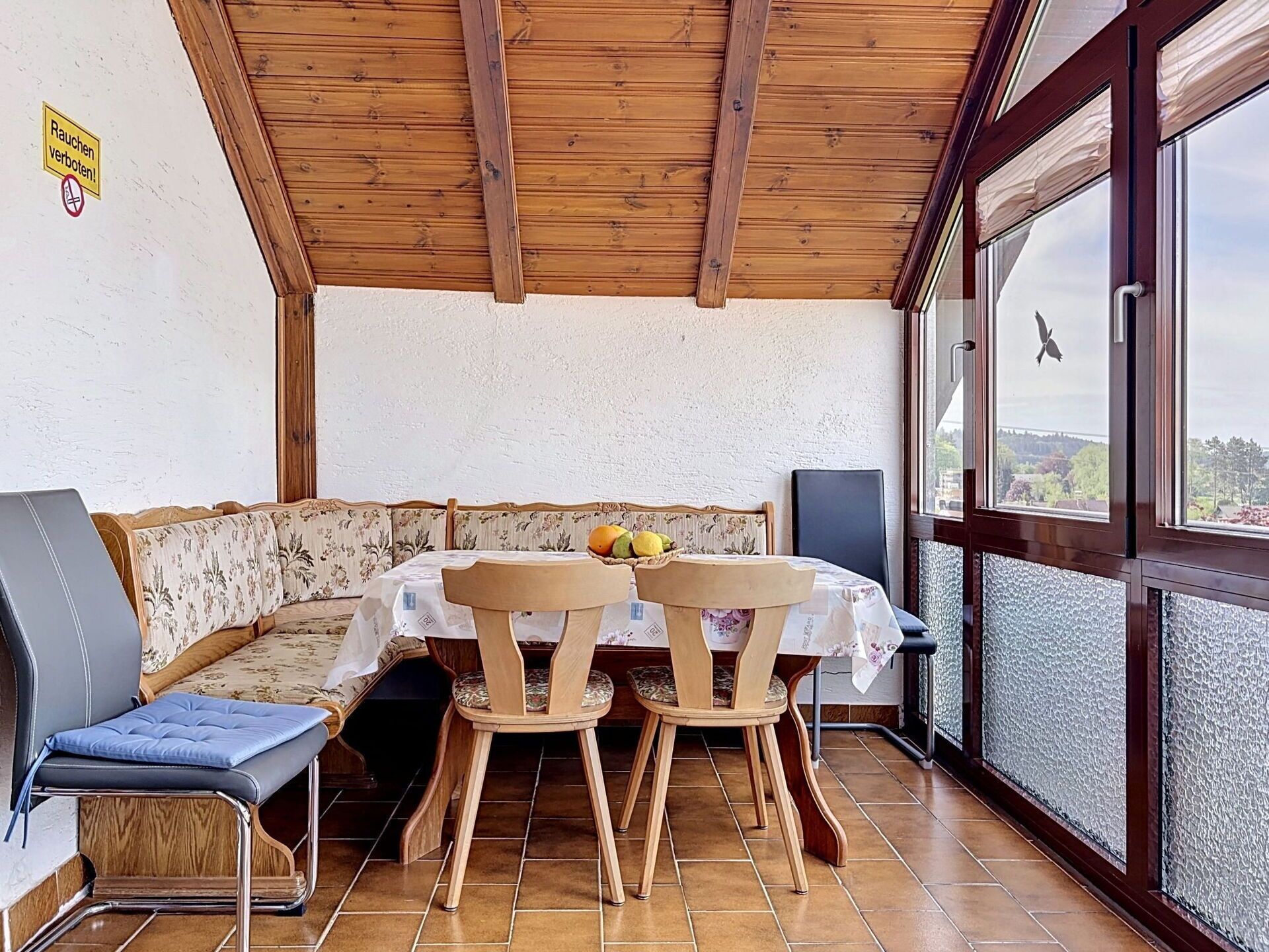 Ein Essbereich mit Holzbalkendecke, einem Tisch mit Stühlen, einer Bank und Blick durch große Fenster.