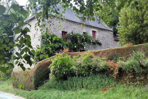 A stone building with red shutters, surrounded by lush greenery and a well-maintained hedge.