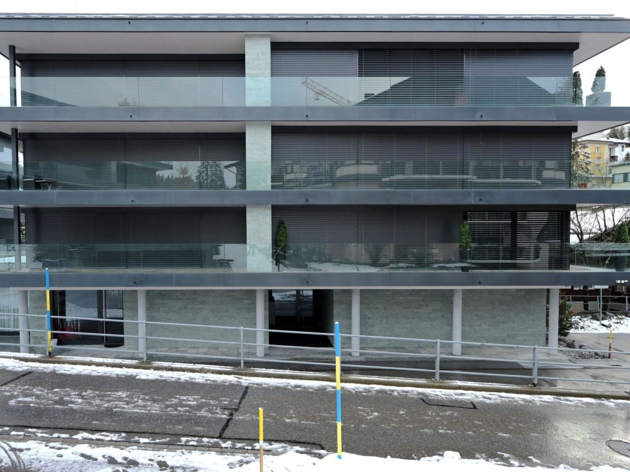 Un edificio de varios pisos con grandes ventanales y balcones, un área de estacionamiento al frente y una grúa de construcción en el fondo.