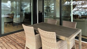 Et moderne udendørs spiseområde med et træbord og fletstole, store skydedøre af glas og udsigt til naturen.