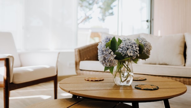 Una mesa de madera redonda con un florero de vidrio transparente que contiene flores, dos posavasos y un sofá blanco al fondo.