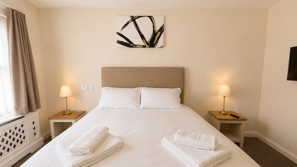 A hotel room with a large bed, two bedside tables with lamps, a wall art piece, and a window with curtains. - Standard Double Room (Banbury)