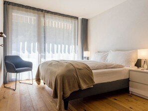 A hotel room with a bed, bedside table, lamp, chair, and large windows with curtains. - Montela Apartment-Hotel & Resort - Holiday Stay Appartement/Fewo by Interhome (Saas-Grund)