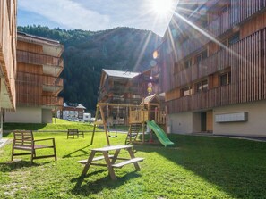 Ein Spielplatz mit Rutsche und Schaukeln, umgeben von Gebäuden mit Balkonen und einem Berg im Hintergrund.