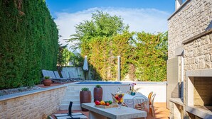 Zona de piscina con barbacoa, sillas de descanso y una mesa con fruta y una bebida.
